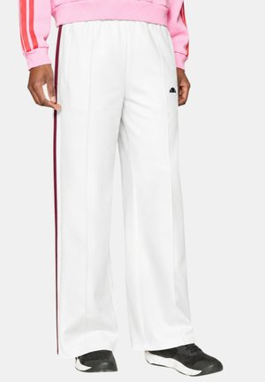 Pantaloni bianchi a gamba larga con strisce laterali rosse e logo nero sulla coscia sinistra, indossati con scarpe da ginnastica nere e bianche e felpa rosa.