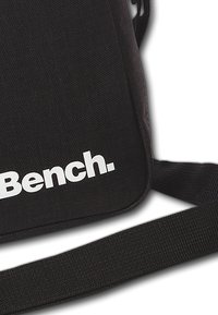 Zwarte stoffen schoudertas met een gladde textuur en een verstelbare schouderband; heeft een opvallend wit "Bench." logo op de voorkant.