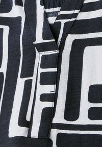 Schwarzer und weißer Stoff mit geometrischen Mustern. Verfügt über einen Reverskragen und kontrastierende Nähte entlang der Kanten. Weiche Textur.