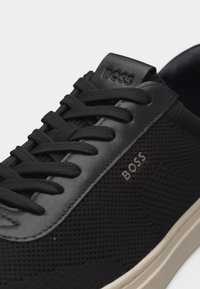 Μαύρο αθλητικό παπούτσι χαμηλού ύψους με υφασμάτινη υφή, δερμάτινη επένδυση, μαύρα κορδόνια, μπεζ σόλα και λογότυπο "BOSS" στο πλάι και τη γλώσσα.