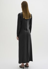 Lange zwarte jurk met lange mouwen, gladde textuur, getailleerde silhouet en een split aan de achterkant. Draagt met zwarte hoge sandalen.