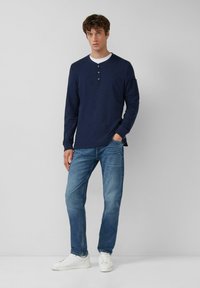 Man in een marineblauwe langslavige henley, lichtblauwe spijkerbroek en witte sneakers, staand met één hand in zijn zak tegen een effen lichte achtergrond.