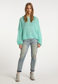 Helle, türkisfarbene, gestrickte Strickjacke mit langen Ärmeln, lässiger Schnitt, kombiniert mit ausgewaschenen, hellblauen Jeans und gemusterten Stiefeletten.