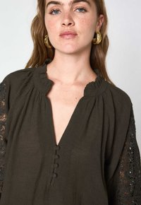Blusa verde oliva scuro con scollatura a volant, dettagli in bottone e maniche in pizzo ricamato. Tessuto leggero e vestibilità comoda.