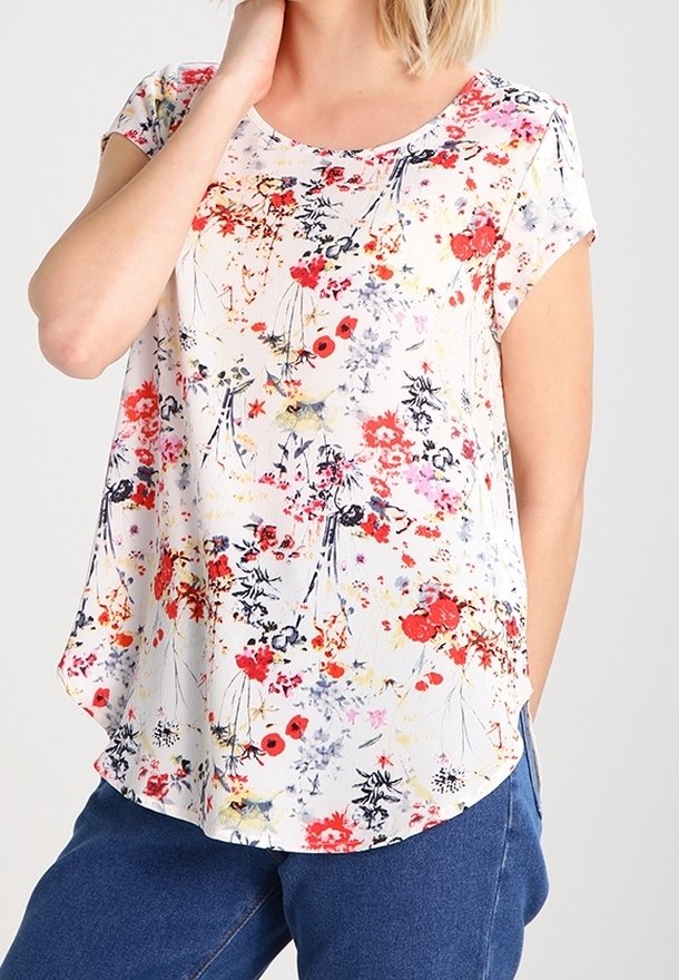 Femme portant un chemisier blanc à manches courtes avec un motif floral rouge, noir et jaune et un jean bleu, main reposant sur le cou.