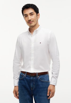 Homme portant une chemise blanche boutonnée et un jean bleu avec une ceinture marron, debout devant un fond blanc uni.