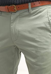 Gröna bomullbyxor med en slät textur, smal passform och sidofickor. Kombinerade med ett brunt läderbälte med ett guldspänne.