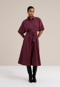 Le modèle pose avec confiance dans une robe bordeaux ceinturée à manches bouffantes, associée à des bottes noires montantes élégantes, le tout sur un fond neutre.