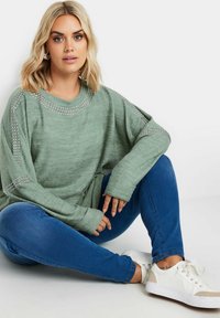 Ljusgrön oversized tröja med silvernitar längs axlarna, i kombination med blå jeans och vita sneakers. Tyget ser mjukt ut.