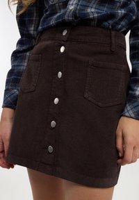 Donna che indossa una gonna di velluto a coste marrone scuro con bottoni frontali e tasche anteriori, abbinata a una camicia a quadri blu a maniche lunghe infilata nella gonna.