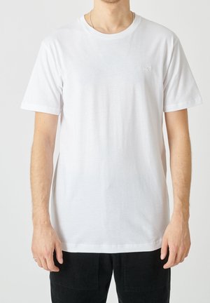 Mężczyzna w prostym białym t-shircie z krótkim rękawem i czarnych spodniach na neutralnym tle, ręce swobodnie opuszczone wzdłuż ciała.