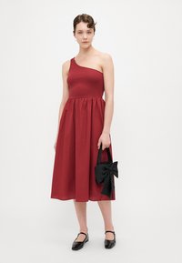 Robe rouge à une épaule avec un haut ajusté et une jupe froncée, accompagnée d'un sac accentué d'un nœud noir. La mannequin porte des chaussures plates noires.