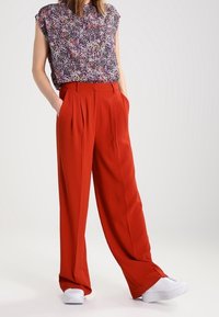 Blusa de estampado floral con mangas de capucha, combinada con pantalones rojos de pierna ancha y cintura alta. El calzado son zapatillas blancas. Tejido suave, corte relajado.