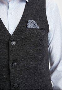 Uomo che indossa un gilet scuro a trama sopra una camicia azzurra a motivo chiaro con un fazzoletto da taschino blu e bianco a motivi.