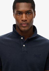 Marineblå polo-shirt med en struktureret overflade, to hvide knapper ved kraven og logo-broderi på brystet. Klassisk design med kort krave.