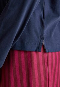 Marineblaues Baumwoll-Oberteil mit langen Ärmeln und einem seitlichen Schlitz, kombiniert mit einem weinrot gestreiften Rock aus strukturiertem Stoff.