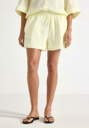 Kvinde iført lysegule teksturerede shorts og matchende top, sorte thongsandaler og et delikat ankelarmbånd, stående på en ensfarvet hvid baggrund.