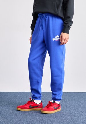 Osoba ubrana w jasnoniebieskie spodnie dresowe Nike Sportswear oraz czerwone buty sportowe Nike z czarnym logo Swoosh, stojąca na niebieskim dywanie.