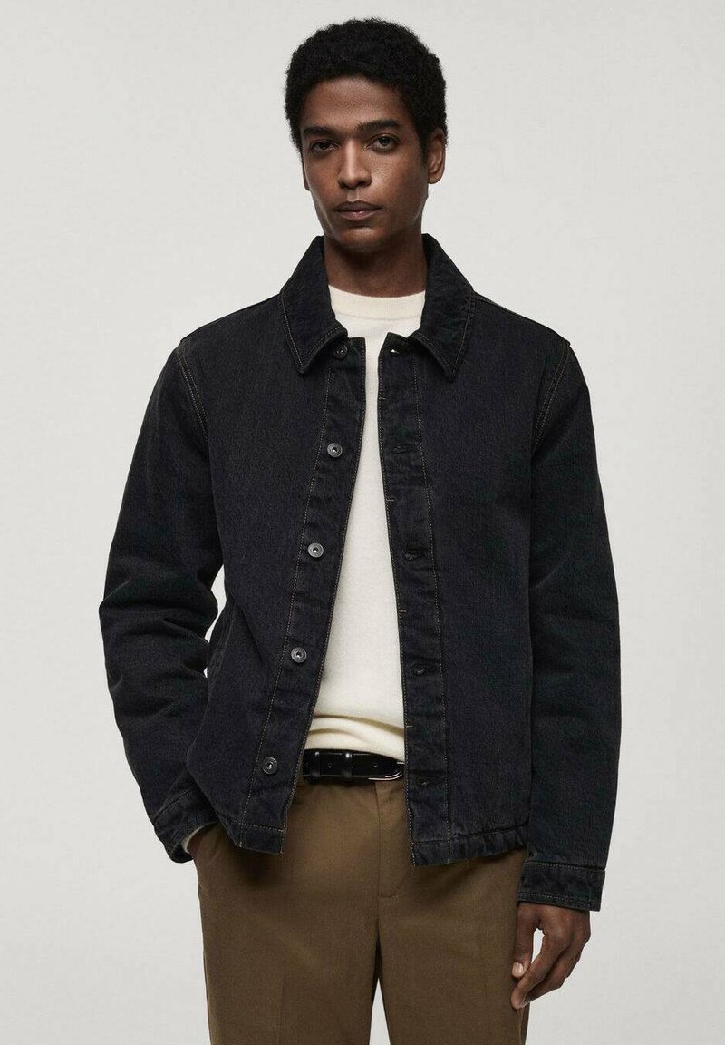 Schwarze Jeansjacke mit Knopfleiste, Umlegekragen und Seitentaschen, getragen über einem hellen Pullover und braunen Hosen. Glatte Textur.