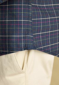 Marineblaues karierte Hemd mit weißen, rosa und grünen Linien, Saumkante sichtbar vor cremefarbenen Hosen mit seitlicher Taschenverzierung.