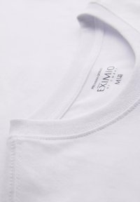 Camiseta de algodón blanca con cuello redondo, que presenta detalles de costura y una etiqueta que indica la marca EXIMIO. Textura suave, diseño minimalista.