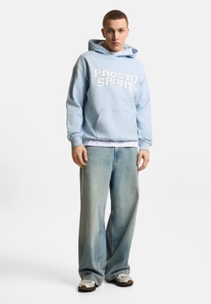 Młody mężczyzna w jasno niebieskiej bluzie z kapturem "Prosto Sport", białym podkoszulku, szerokich jasnych dżinsach i białych tenisówkach stoi na białym tle.
