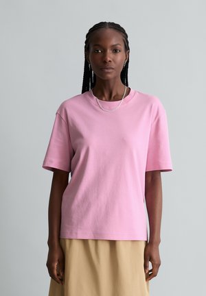 Woman with braided hair wearing a light pink short-sleeve t-shirt and beige skirt standing against a plain gray background.