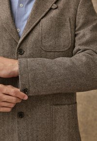 Brown herringbone blazer with a structured fit, featuring a patch pocket and buttoned cuffs. Textured fabric with a subtle sheen.