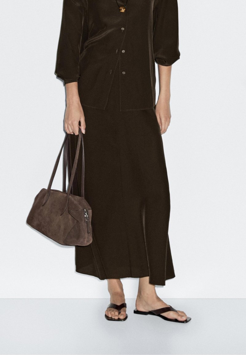 Bruine blouse met knopen en bijpassende lange rok van zachte stof. Houdt een donkerbruine suède handtas met dubbele handvatten vast. Zwarte slippers.