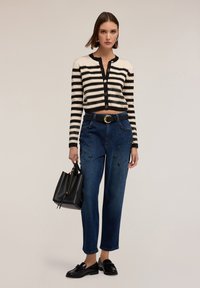 Cárdigan recortado a rayas en blanco y negro, jeans azul adornados, cinturón negro con hebilla dorada, mocasines negros, sosteniendo un bolso negro.