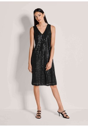 Frau in einem ärmellosen, schwarzen Pailletten-Kleid bis zum Knie und schwarzen Riemchen-Absätzen, stehend vor einem neutralen Hintergrund.