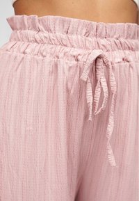 Un pantalon texturé de couleur rose clair avec une taille smockée et un détail de lien. Le tissu a un aspect froissé, ce qui renforce son allure décontractée.