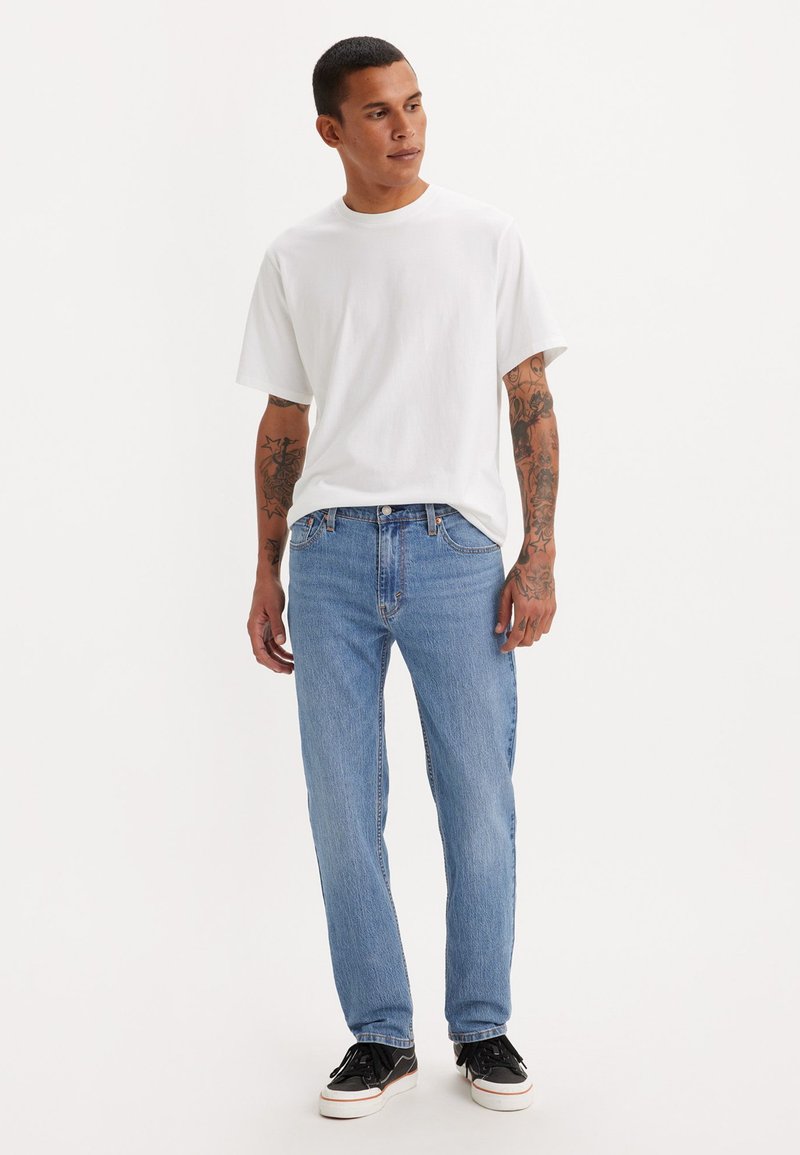 Camiseta blanca de manga corta, jeans rectos azul claro, zapatillas negras con suelas blancas. Tatuajes visibles en los brazos. Conjunto simple y casual.