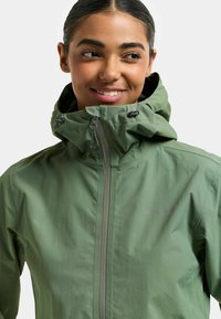 Grüne wasserdichte Jacke mit hohem Kragen, verstellbarer Kapuze und Frontreißverschluss. Leichtes Material mit dezentem Schimmer und Logoakzent.