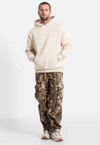 Sudadera de color beige con bolsillo frontal y capucha con cordón, combinada con pantalones cargo de camuflaje en tonos marrón y beige, que cuentan con bolsillos laterales.