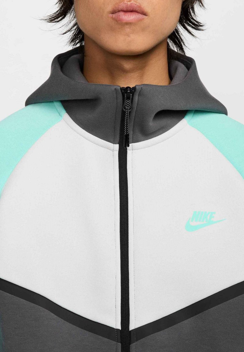 Sudadera con cremallera de Nike que presenta un cuerpo gris con acentos en aqua, una cremallera negra y un pequeño logo de Nike en aqua en el pecho. Textura suave, ajuste atlético.