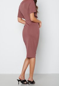 Robe midi mauve en tissu extensible, avec des manches courtes, une silhouette ajustée et une longueur de genou. Sandales à talons noires avec des accents.