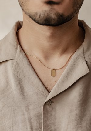 Goldene Halskette mit rechteckigem Anhänger, getragen über einem beigen Hemd mit Kragen aus leichtem, strukturiertem Stoff.