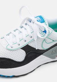 Sneaker bianco in rete con accenti in pelle scamosciata nera, dettagli turchesi e suola grigia. Presenta lacci bianchi e un'etichetta con il logo sulla linguetta.