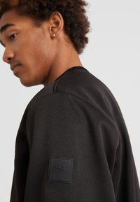 Zwarte sweatshirt met een gestructureerde stof, een ronde halslijn en een rubberen logo-patch op de bovenarm.