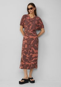 Maxi jurk in aardetinten met een abstract bruin en roest patroon, korte pofmouwen, knopenfront en een zijsplit aan de onderrand. Zwarte platform sandalen.