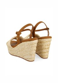 Sandalias de cuña de cuero marrón con detalles de paja trenzada, que cuentan con correas ajustables en los tobillos y un diseño abierto redondeado. Apariencia natural texturizada.