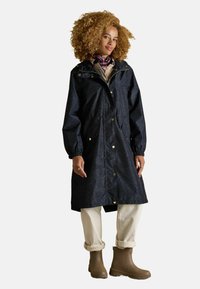 Trench coat blu navy impermeabile con chiusura frontale a zip, accessori dorati, polsini elasticizzati e due tasche laterali. Abbinato a pantaloni beige e stivali marroni.