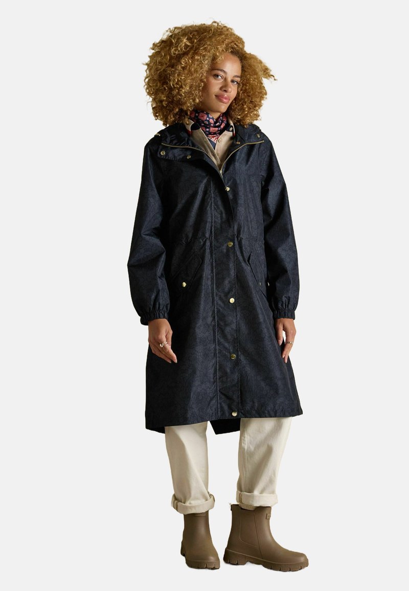 Trench coat blu navy impermeabile con chiusura frontale a zip, accessori dorati, polsini elasticizzati e due tasche laterali. Abbinato a pantaloni beige e stivali marroni.