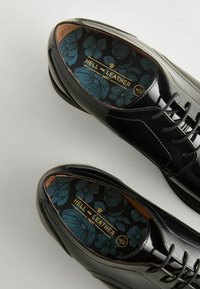 Schwarze Lacklederschuhe mit glatter Oberfläche, ausgestattet mit einer blumenmusternden Innensohle in Teal und goldener Beschriftung sowie traditionellem Schnürdesign.