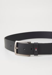 Ceinture en cuir noir avec une boucle en métal argenté, ornée d'un accent décoratif avec un petit logo rouge, blanc et bleu près de la boucle.