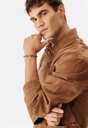 Joven con chaqueta marrón apoyando la barbilla en la mano, lleva una pulsera de cuentas, mirando pensativamente a la cámara sobre un fondo blanco.