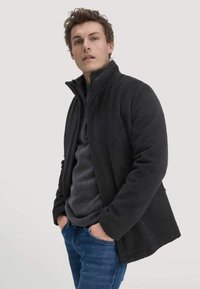 Schwarze Fleecejacke mit hohem Kragen, Reißverschluss und strukturierter Stoff; getragen über einem grauen Pullover, kombiniert mit blauen Jeans.