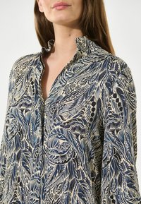 Camicia a stampa floreale in crema, blu e nero. Presenta un colletto alto e chiusura a bottoni. Il tessuto ha una finitura strutturata con pattern intricati.