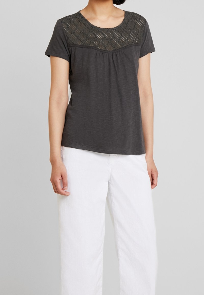 Haut gris à manches courtes avec un empiècement en dentelle texturée, encolure dégagée et coupe ample, associé à un pantalon blanc à jambes larges.